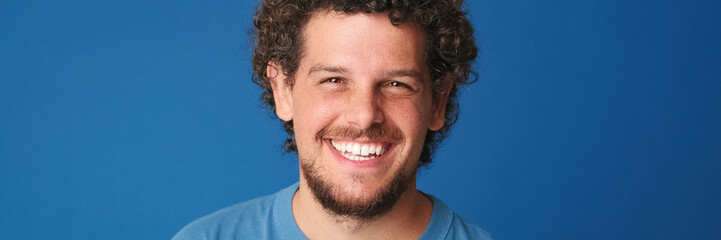 Fototapeta premium Close up, guy with curly hair dressed in blue t-shirt looks at the camera with smile isolated on blue background in the studio. Panoramic. Panoramic