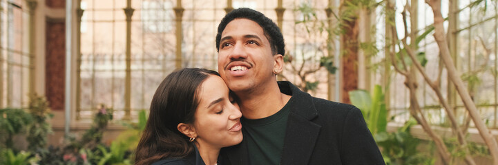 Fototapeta premium Happy young couple embracing in greenhouse, with background of arched windows and plants. Panoramic
