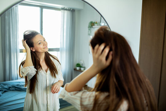 Smiling young woman fixing hair in bedroom mirror