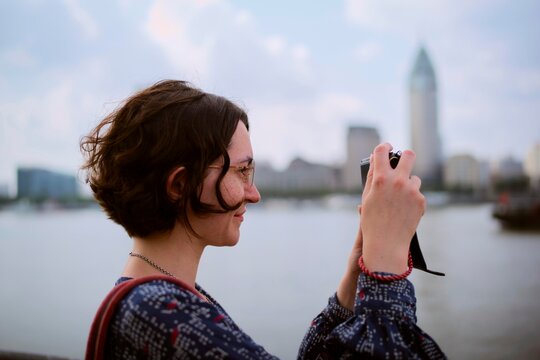 Woman Takes Picture by River in City With Buildings in Background