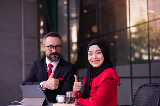 Two business professionals, including a Muslim woman wearing a Islam hijab, discussing work on a tablet at an outdoor caf&eacute;.