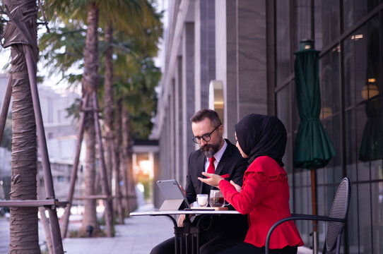 Two business professionals, including a Muslim woman wearing a Islam hijab, discussing work on a tablet at an outdoor caf&eacute;.