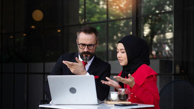Two business professionals, including a Muslim woman wearing a Islam hijab, discussing work on a tablet at an outdoor caf&eacute;.