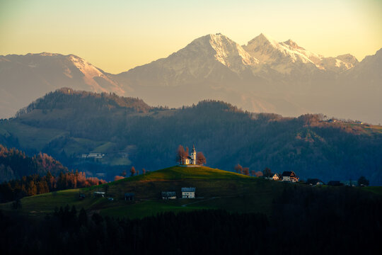 Aerial view of the historic Saint Thomas Church on a green hilltop with the snow-capped Julian Alps in the background during golden hour Slovenia.
