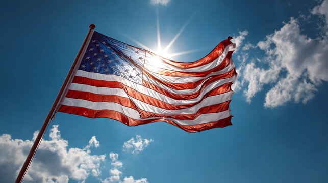 The American flag is backlit by the sun, creating a dramatic silhouette against a cloudy blue sky.