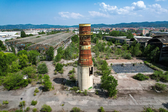 Aerial view of an abandoned industrial site featuring a rusty smokestack and dilapidated factory buildings overgrown with vegetation Settimo Torinese, Piedmont, Italy.