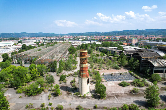 Aerial view of an abandoned industrial site featuring a tall rusty chimney and derelict warehouses surrounded by overgrown vegetation in Settimo Torinese, Piedmont, Italy.