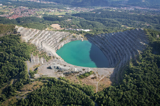 Aerial view of the Amiantifera di Balangero open-pit mine featuring a turquoise lake surrounded by terraced rock walls and lush green forests in Balangero, Piedmont, Italy.