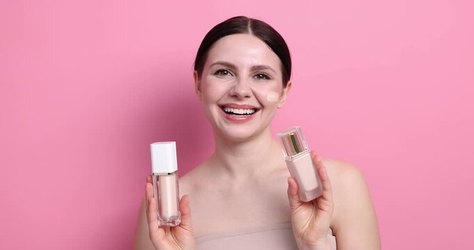 Beautiful woman with bottles of foundation and smear on her face against pink background