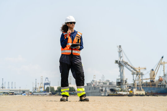 Full body of female engineer in safety vest and helmet using tablet and radio at shipyard port for logistics and industrial operation.