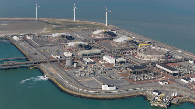 Aerial view of the Fluxys LNG Terminal Zeebrugge featuring large circular storage tanks, industrial infrastructure, and wind turbines along the coast in Brugge, Vlaanderen, Belgium.