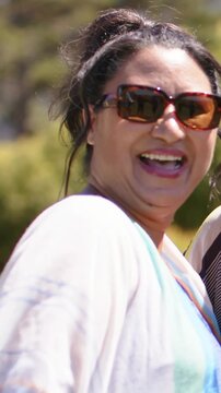 Vertical video: Laughing woman with coverup, spotting bee, waving to camera, friend joining in park