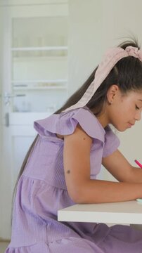 Vertical video: Writing student leaning forward at kitchen nook in lavender dress using pink pencil