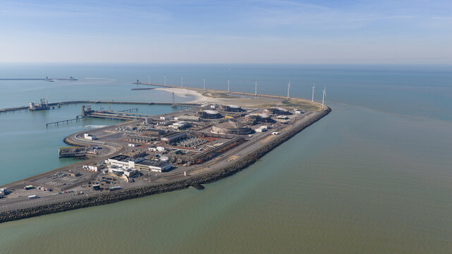 Aerial view of Fluxys LNG Terminal Zeebrugge with large storage tanks and wind turbines along the coastline under a clear blue sky in Brugge, Vlaanderen, Belgium.