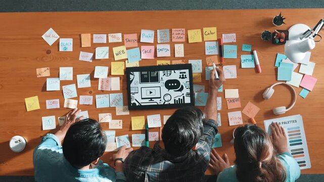 Top view of creative designer working together brainstorming idea by using sticky notes at meeting table with color palettes. Aerial view of smart team choosing icon graphic from tablet. Symposium.