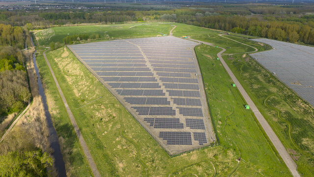 Aerial view of the Zonnepark Braambergen solar farm featuring rows of photovoltaic panels on a green hillside surrounded by lush trees and a narrow canal in Almere, Flevoland, Netherlands.
