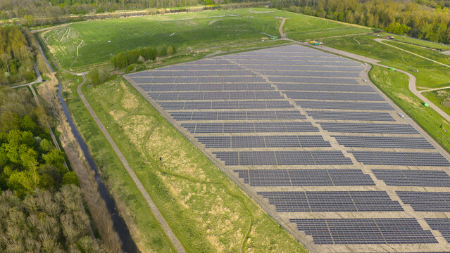 Aerial view of Zonnepark Braambergen solar farm with rows of photovoltaic panels on a green slope surrounded by lush forest and a canal in Almere, Flevoland, Netherlands.