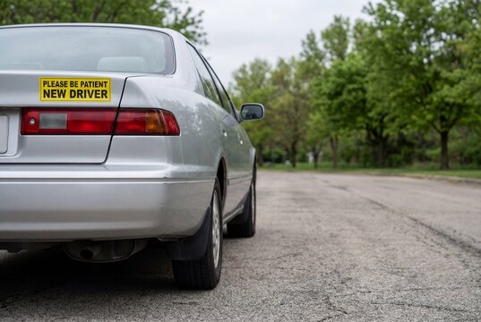 Silver sedan is displaying a yellow bumper sticker that reads Please Be Patient New Driver.