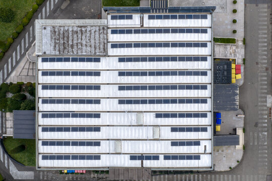 Aerial view of rows of solar panels installed on the white roof of an industrial building surrounded by paved streets and green bushes Reggello, Tuscany, Italy.