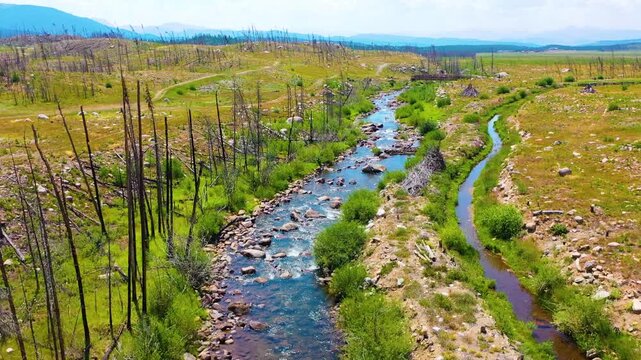 Cinematic backward moving drone view revealing a clear creek across alpine meadowland with stone lined water sparse trees sunlit grass open valley terrain and hazy peaks beyond