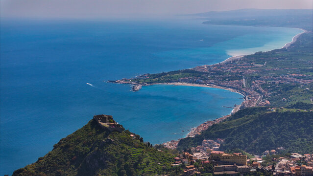 Aerial view of the Saracen Castle on a hilltop overlooking the turquoise waters of Giardini Naxos bay and the curved coastline of Taormina, Sicily, Italy.