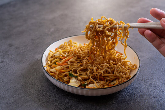 Javanese fried noodles or Bakmi Jawa lifted with chopsticks from bowl on dark background, traditional Indonesian noodle dish with vegetables and savory flavor
