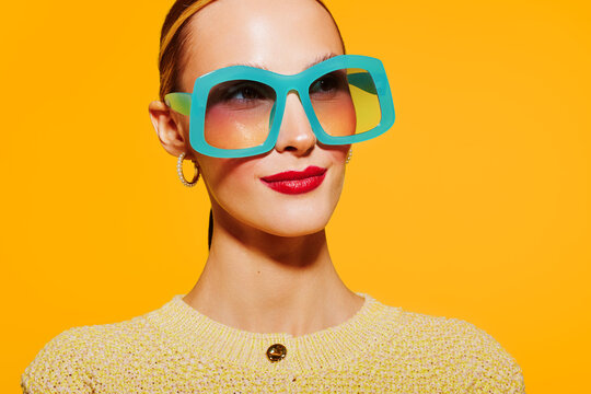 Stylish woman with blue oversized sunglasses and red lipstick poses against bright yellow background. Fashion portrait with vibrant colors and confident expression in studio lighting.
