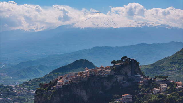Aerial view of the historic hilltop village of Castelmola perched on a rocky cliff with the snow-capped Mount Etna and soft clouds in the background in Castelmola, Sicily, Italy.