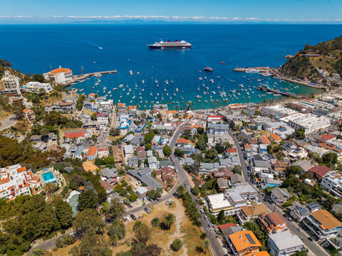 Aerial view of a coastal town with a busy harbor full of boats, winding streets, and a large cruise ship anchored offshore.