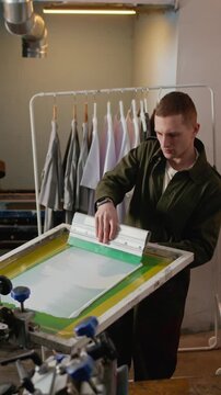 Vertical medium shot of worker dragging squeegee to spread white ink evenly across mounted printing screen