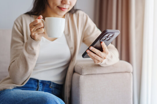 Young Asian woman drinking coffee and using mobile phone on a sofa at home in the morning