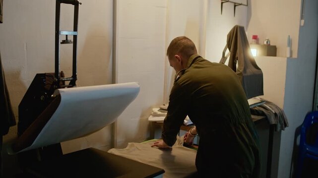 Side view medium shot of man organizing and folding printed t shirt on table in workshop, showing apparel preparation and product handling