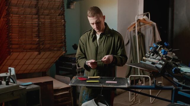 Medium stab shot of young Caucasian male professional in coverall sorting colorful palettes for screen printing and branding jobs inside workshop environment filled with machinery equipment