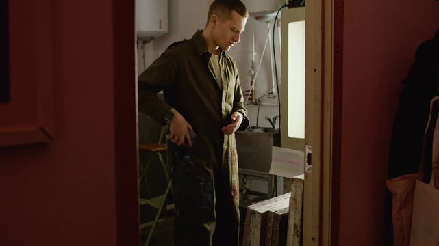 Wide shot of Caucasian male worker selecting and organizing screen printing frames on rack in workshop after using his smartphone during minute break from branding work