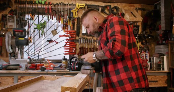 Sanding wood. Carpentry tool. Male joiner in red red plaid shirt smoothing plank surface with professional electric equipment in workplace.
