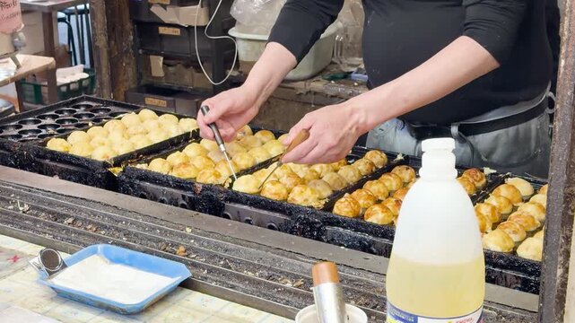Skilled street vendors in Japan prepare takoyaki, golden-brown octopus balls, on a hot griddle with dynamic motion.