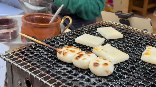 A close up video of dango being grilled on charcoal at a food stall during a Japanese festival or market with steam rising from a clay pot in the background.