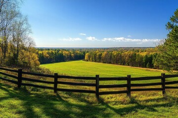 Fototapeta premium wooden fence overlooking sunlit green meadow and autumn forest under a clear blue sky, peaceful countryside panorama