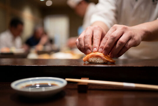 Close-up of chef hands shaping fresh salmon nigiri sushi on a wooden counter with chopsticks and soy sauce dish in a blurred restaurant setting, showing traditional sushi preparation and kitchen actio