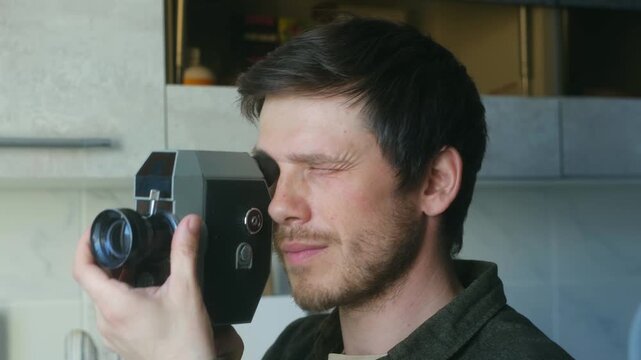 Young cameraman with a beard holding an old fashioned camera, looking through the viewfinder and squinting with concentration while capturing vintage style footage for a film project