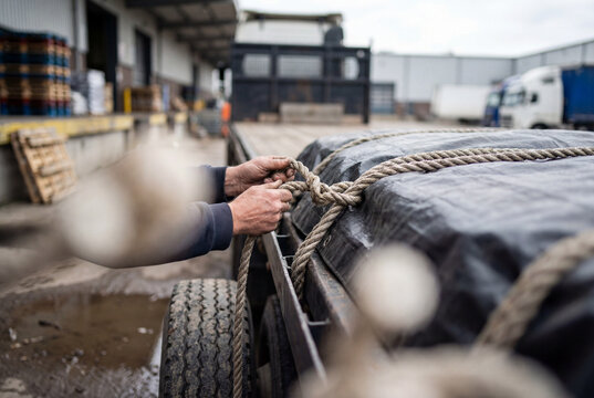 Close-up of hands tying rope securing covered cargo on flatbed truck at industrial loading dock with warehouse and other trucks in background