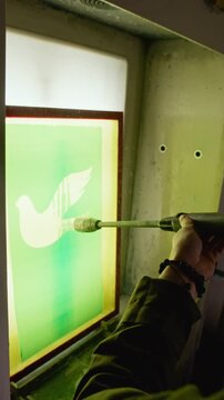 Vertical shot of worker applying coating with spray gun to screen printing frame in dimly lit workshop, capturing professional print production process as bird design being formed