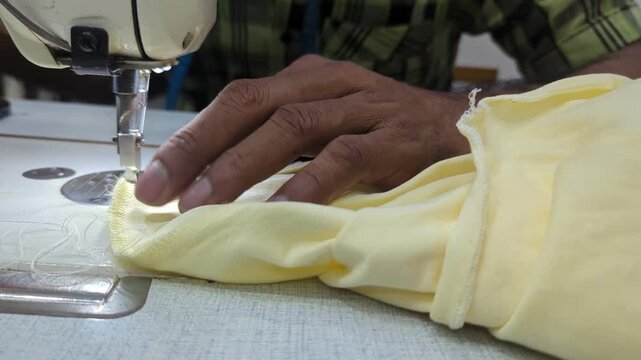 Close-up of professional seamstress hands operating an industrial sewing machine to stitch the hem of a yellow cotton garment in a textile factory.