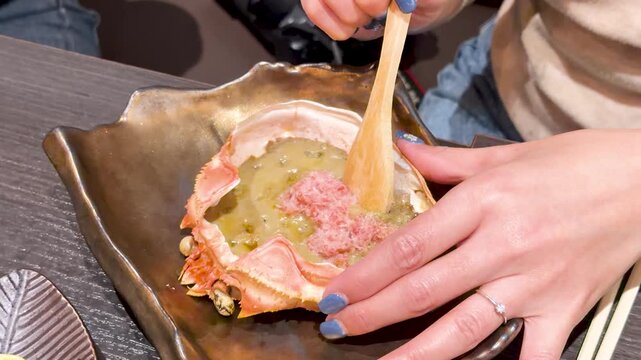 Woman hands mixing delicious japanese kani miso crab roe inside shell with wooden spoon in restaurant.