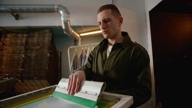 Eye level shot of male worker spreading white ink with squeegee over screen printing frame mounted on machine, showing detailed printing workflow