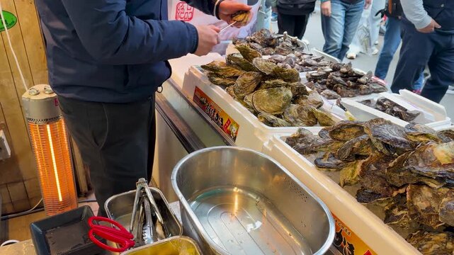 Vendor shucking fresh oysters on ice at busy outdoor market stall footage.