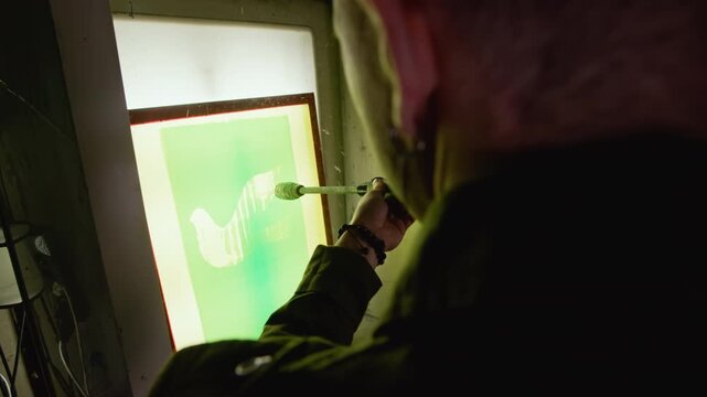 Over shoulder closeup shot of worker using spray gun on screen printing frame in low lit workshop during professional print production and coating process
