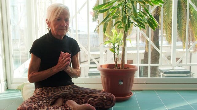 Senior woman with white hair meditating in the lotus position with hands in prayer on a bright balcony with a large potted plant and Mediterranean background. Perfect for wellness aging concept