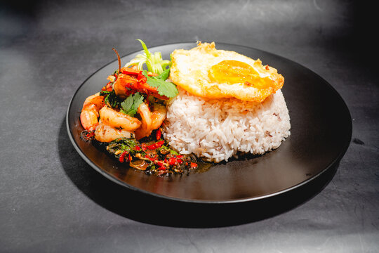 Stir-fried shrimp with holy basil served over rice and topped with a fried egg on a black plate.
