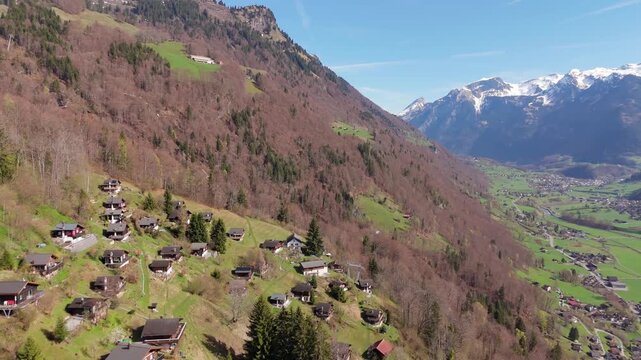 Alpine hamlet of Schlattberg above Luchsingen with traditional chalets on a hillside and the Glarus valley in the distance. Snow-capped mountains visible in background. Aerial drone shot.